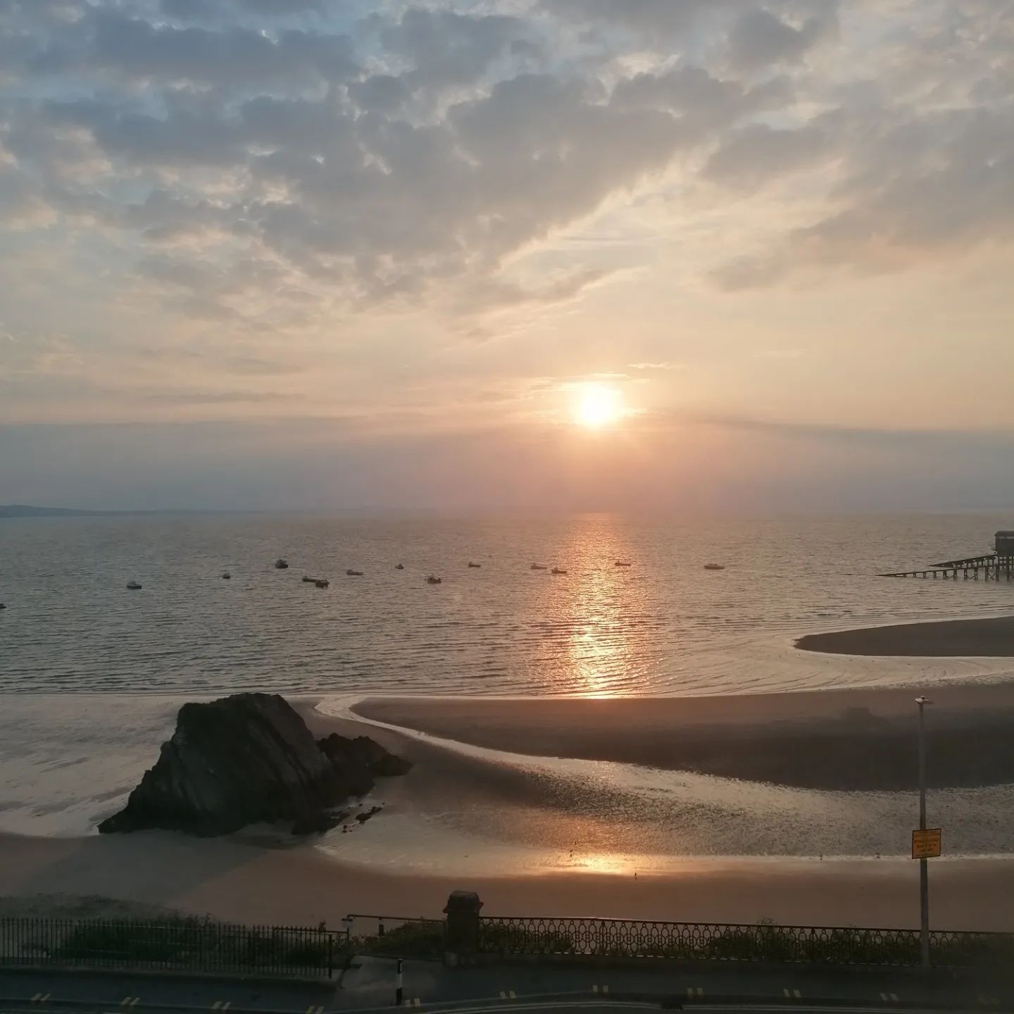 Sunset over ocean, beach in foreground, rock formations, boats in water, cloudy sky, warm colors.