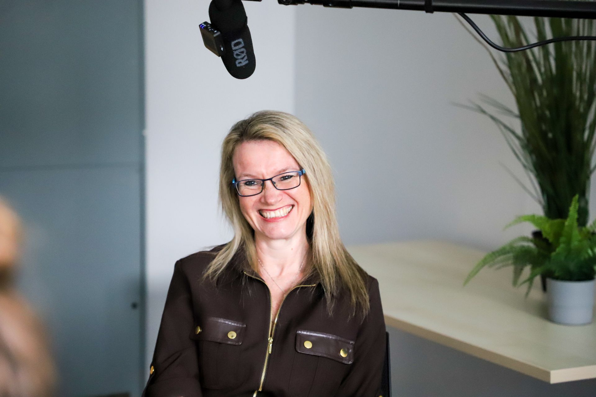 Woman in glasses smiling, wearing a brown jacket, with a microphone overhead and plant in the background.