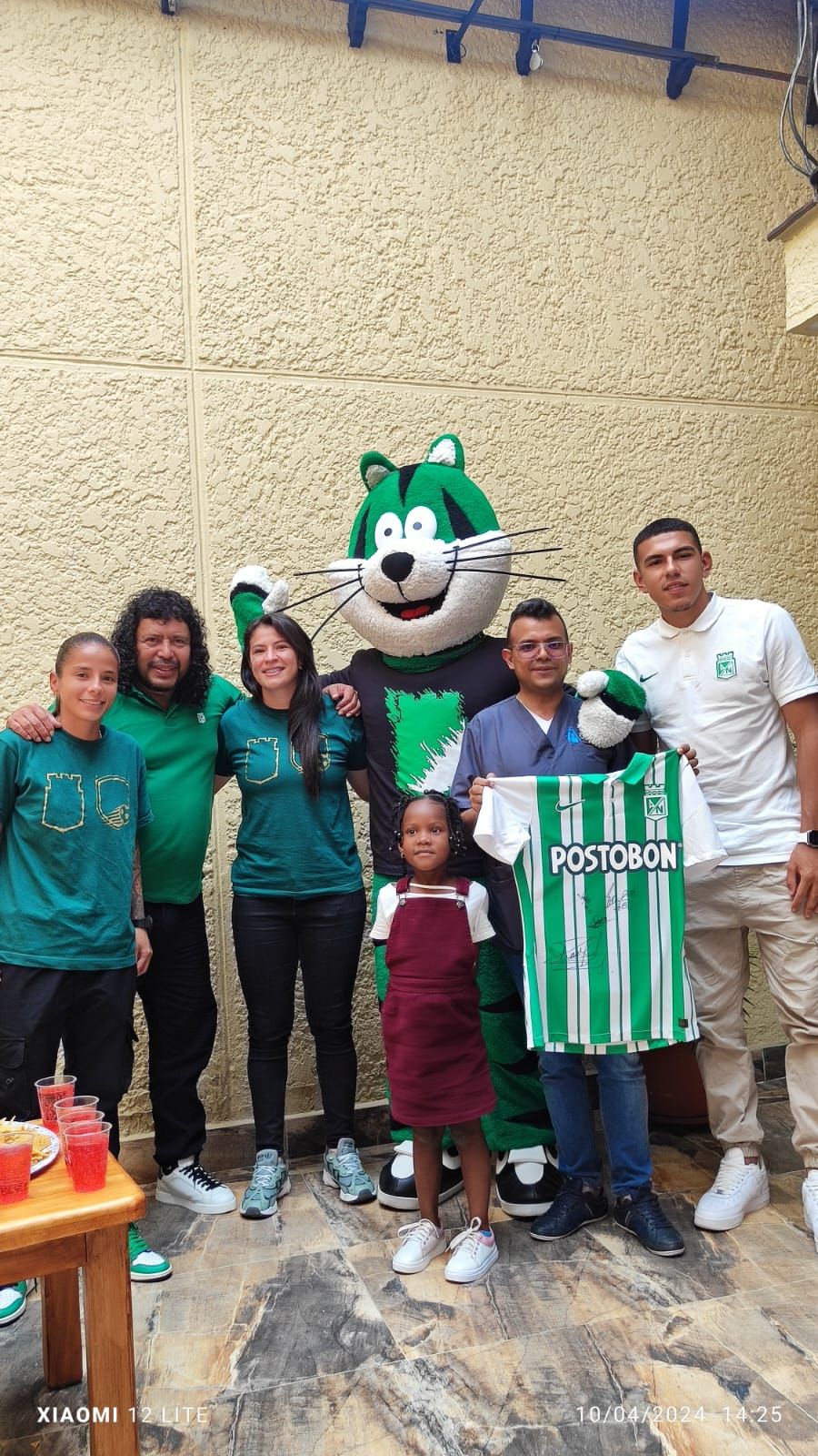 Un grupo posa con una mascota que sostiene una camiseta de fútbol, vestida de verde y blanco