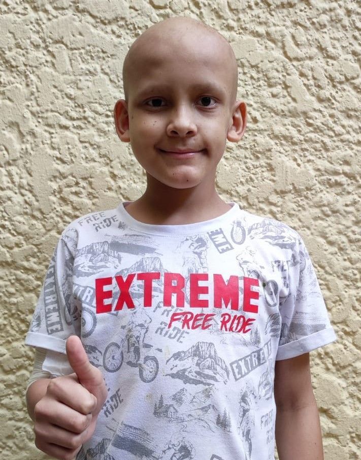 Niño con la cabeza rapada, sonriendo, levantando el pulgar. Con camiseta blanca y la palabra