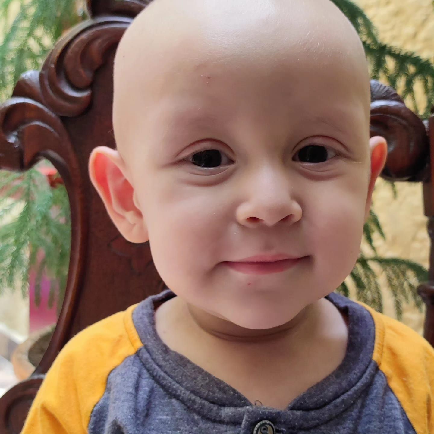Niño calvo sonriente con una camisa amarilla y gris, sentado en el interior.