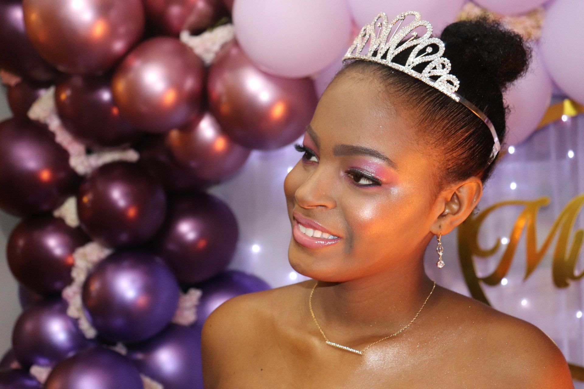 Mujer con tiara y sombra de ojos rosa sonríe, posando frente a globos morados.