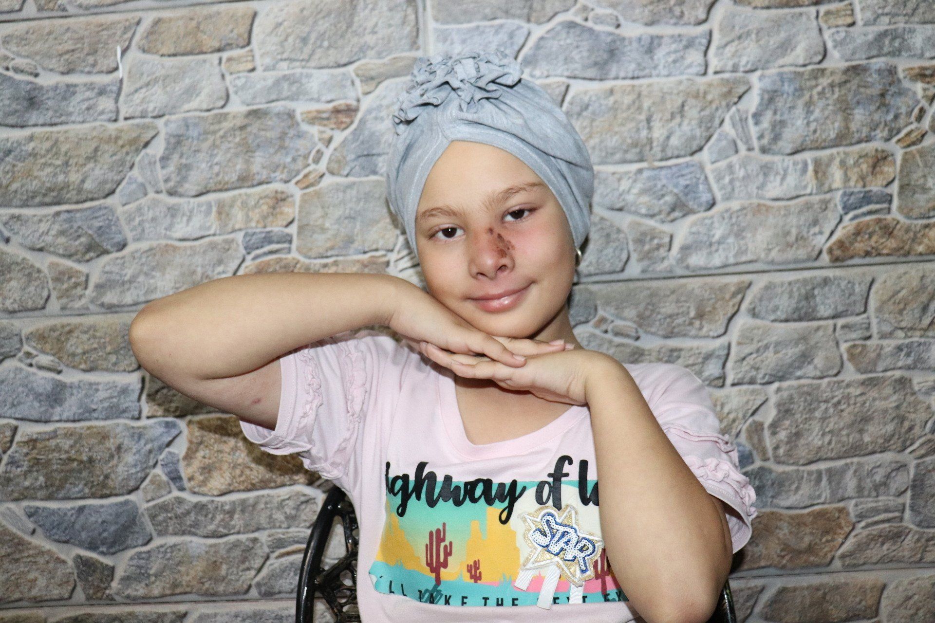 Una niña con una cubierta azul claro en la cabeza y una camisa rosa posa con las manos cerca de la cara 