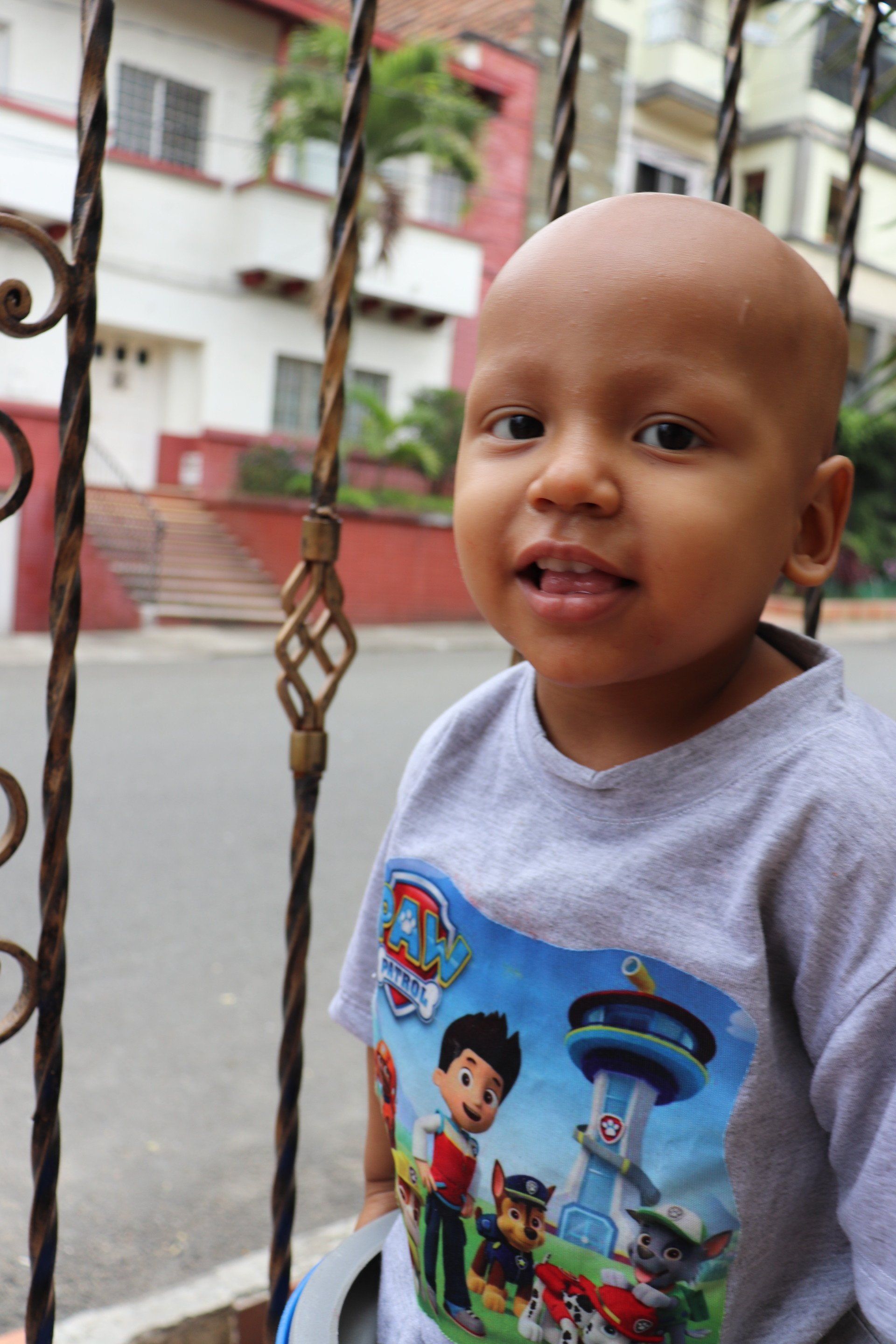 Un niño calvo que viste una camiseta de Paw Patrol sonríe, parado cerca de una puerta de metal 