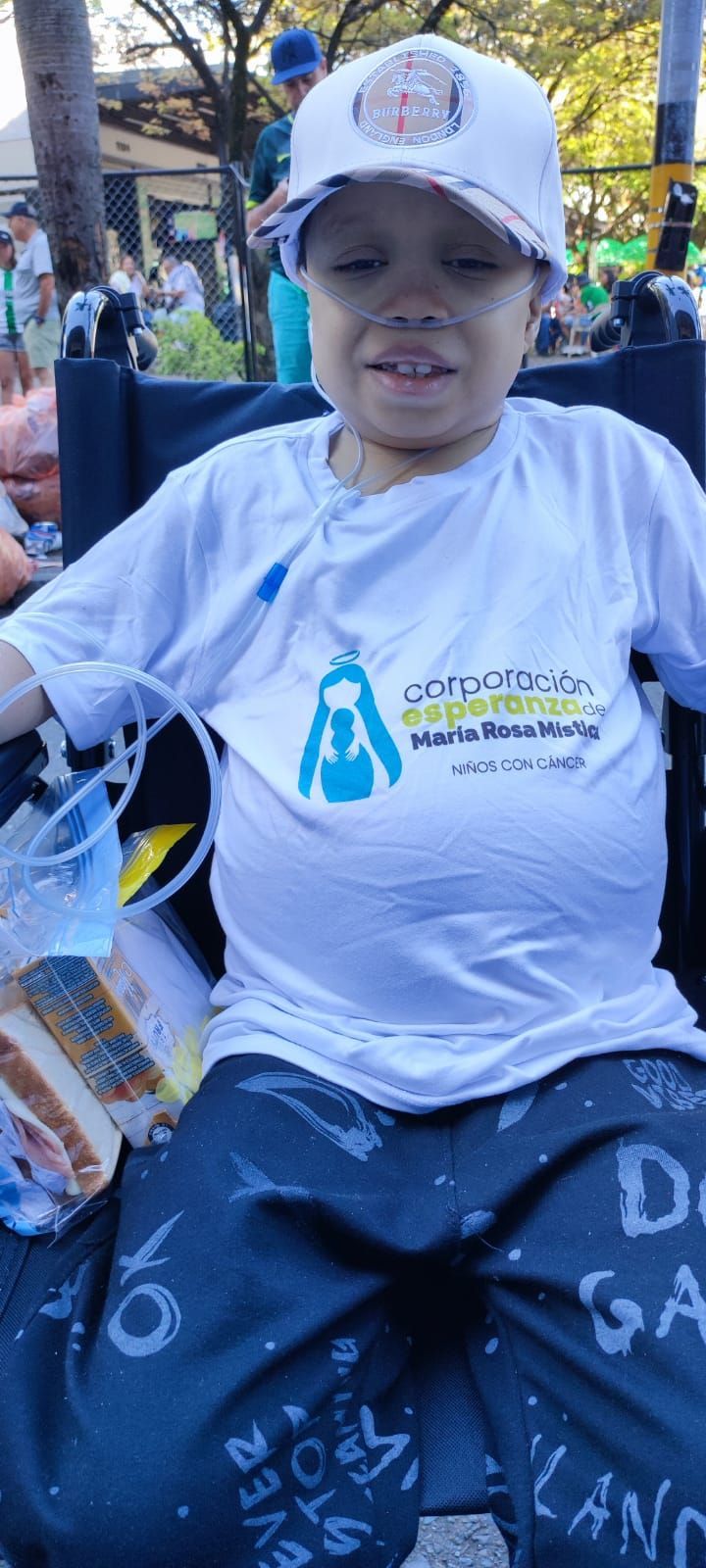 Una persona en silla de ruedas lleva gorra, camisa blanca con el logo y pantalones azules. Sonríe.