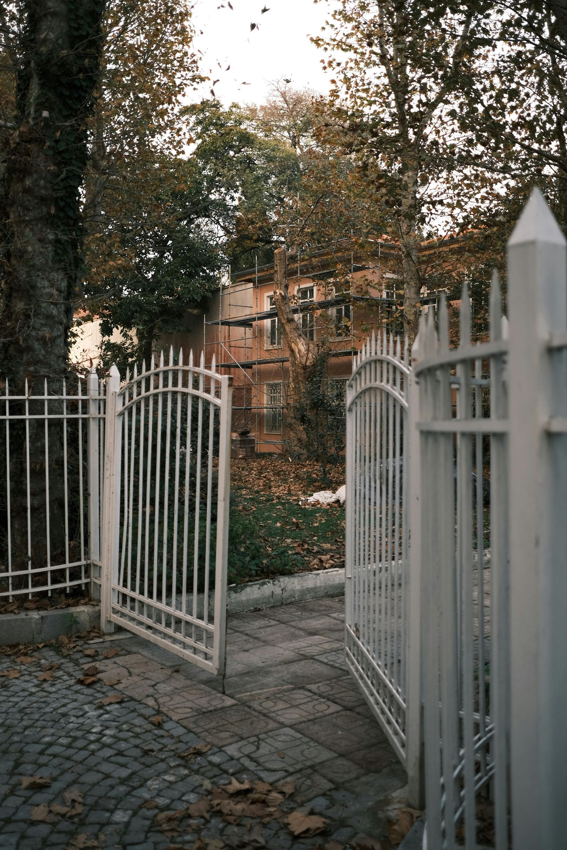 Open white gate beside a cobblestone path, leading to a house and trees in a yard.