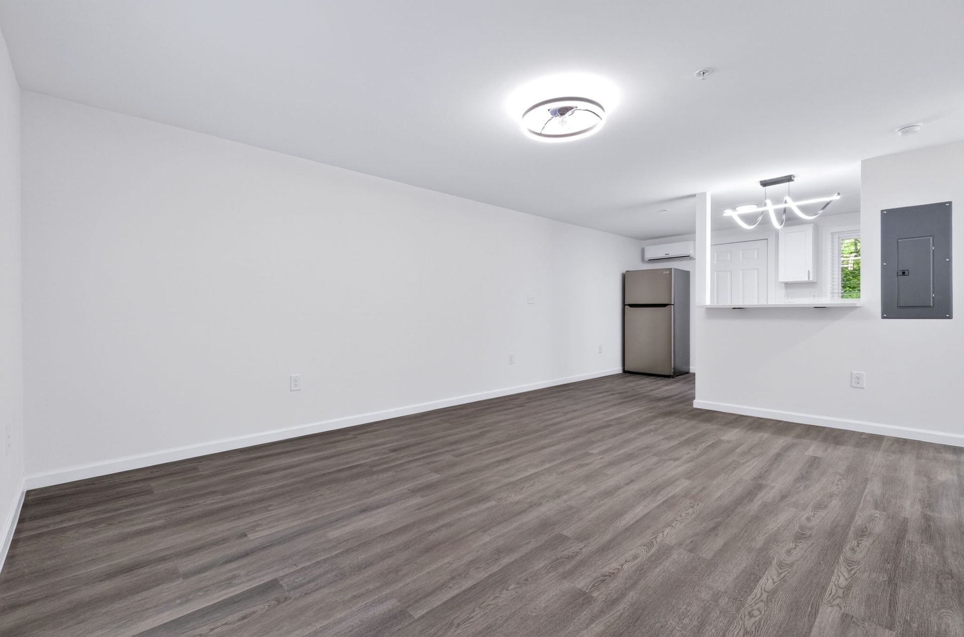 Empty white room with gray carpet, ceiling light, and small kitchen area in the back right