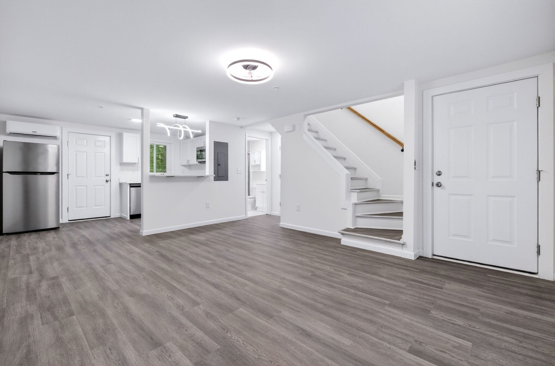 Bright empty living room with gray floors, white walls, stairs, kitchen, and stainless steel refrigerator