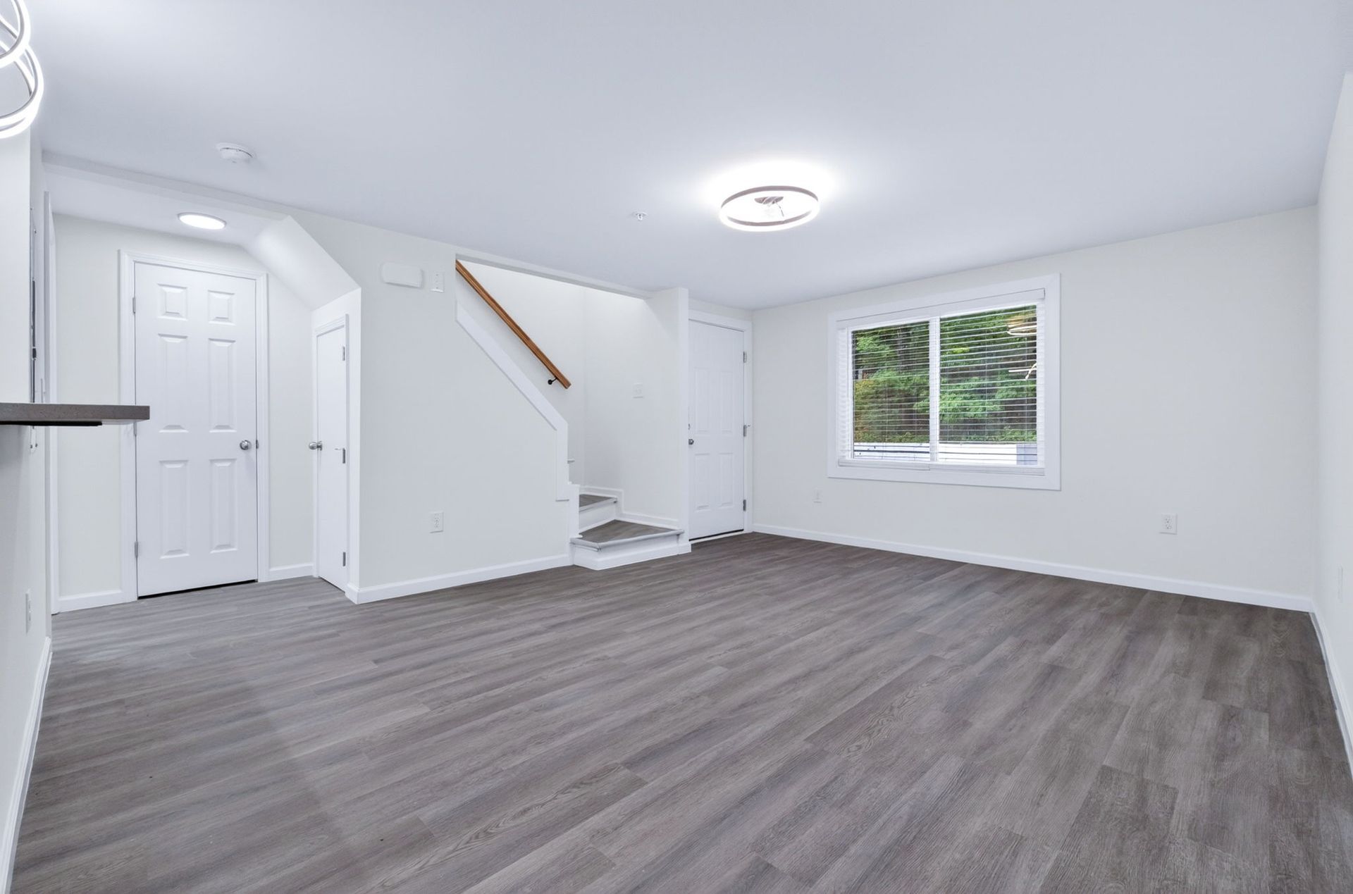 Empty white living room with gray flooring, staircase, front closet, and window.