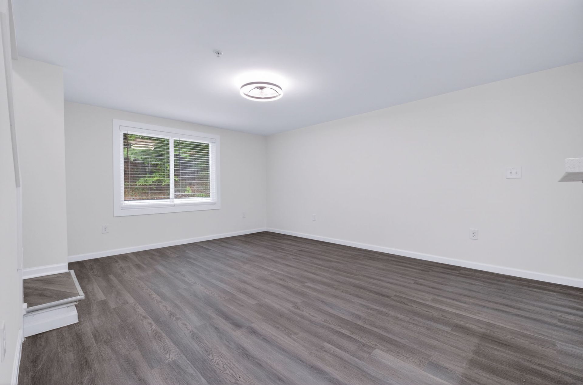 Empty white room with gray wood-look flooring, ceiling light, and a small window.