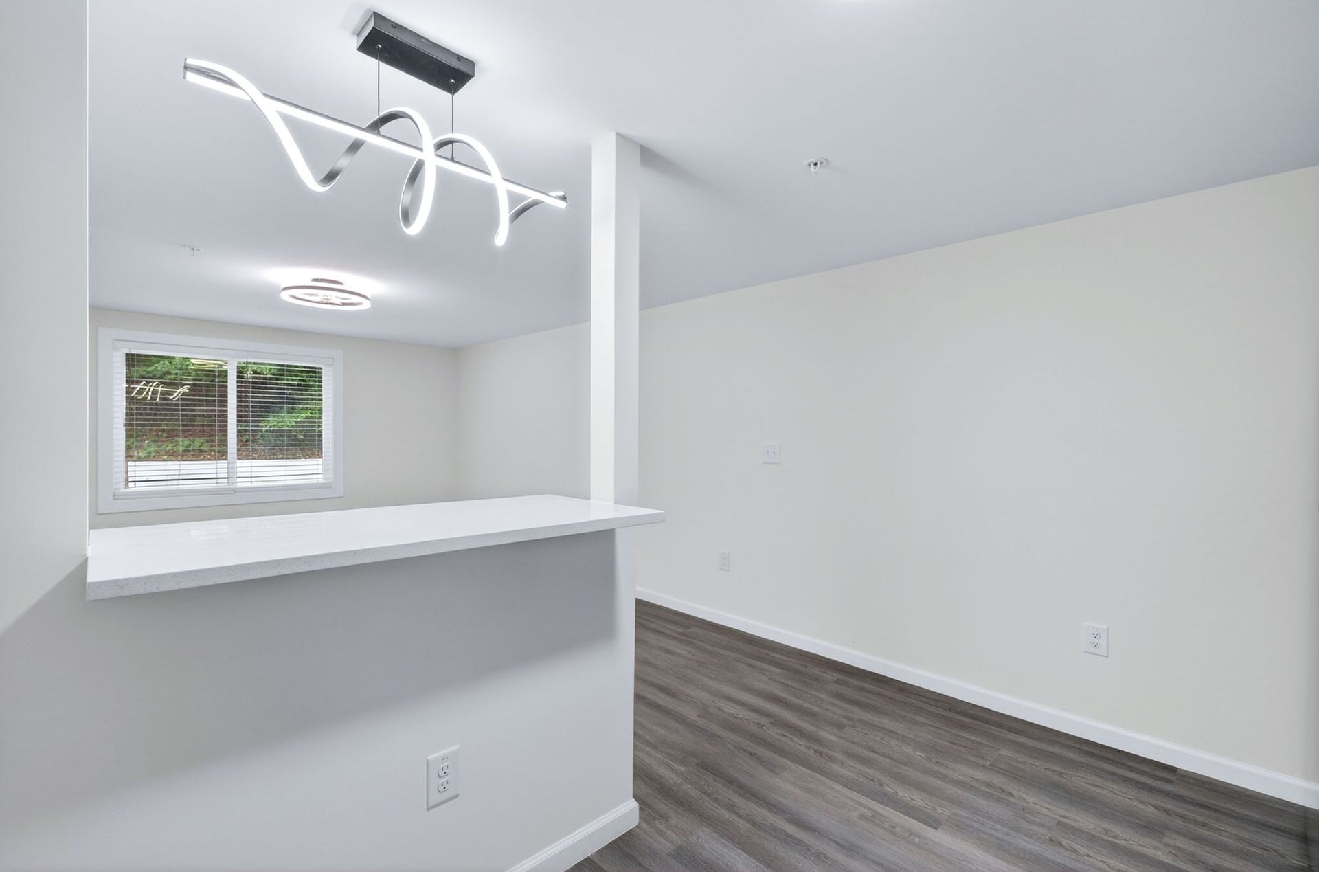 Empty modern room with white walls, wood floor, and a pass-through counter under a contemporary light fixture
