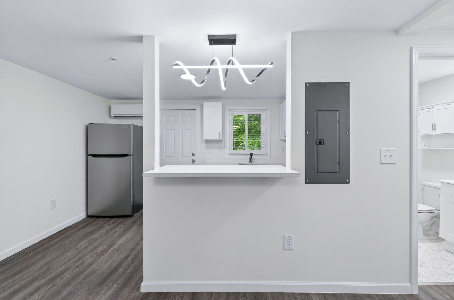 Modern open kitchen with stainless fridge, white counters, and dark wood flooring leading to a hallway.