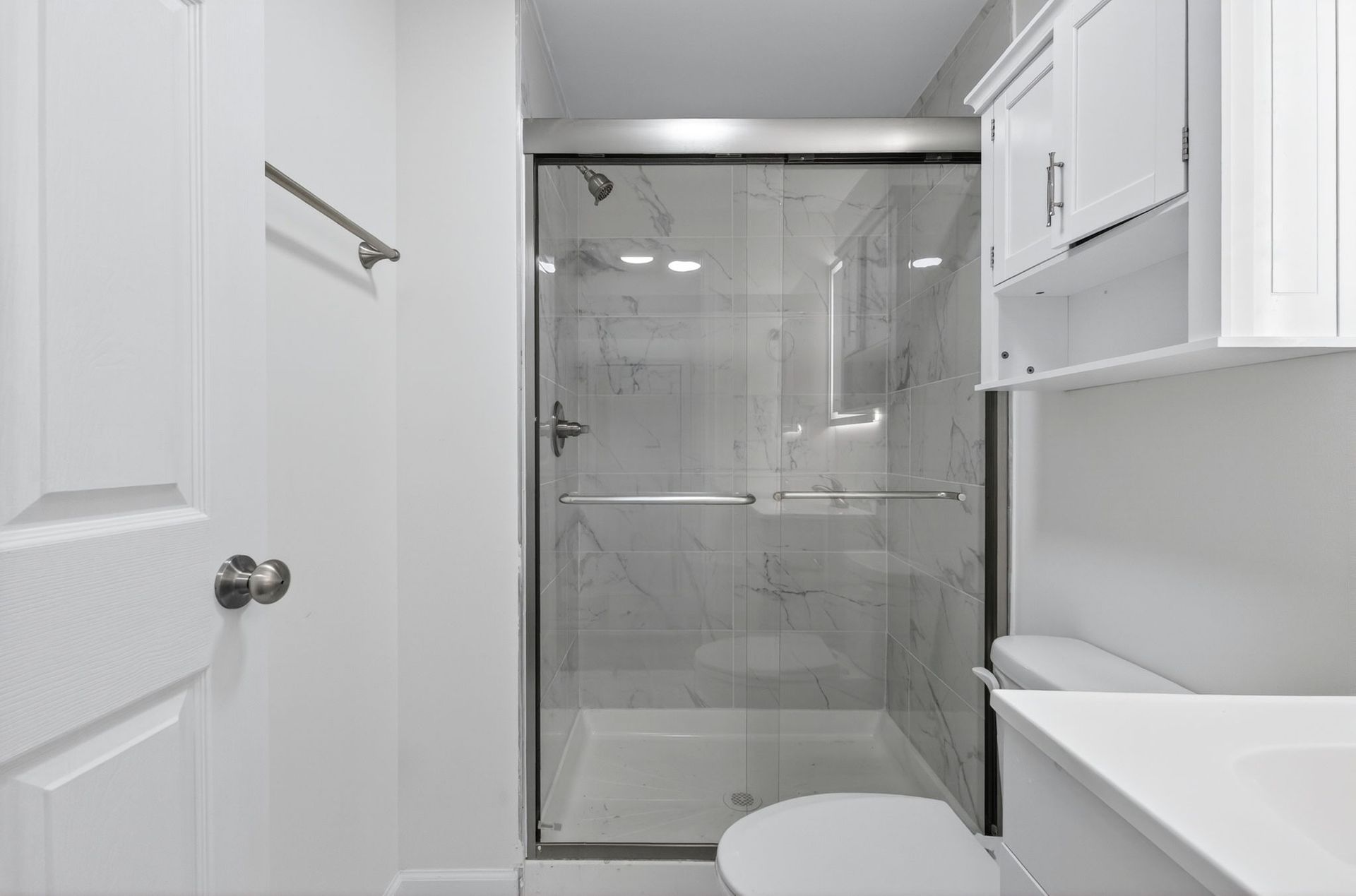White bathroom with shower, toilet, and vanity in a narrow, modern space