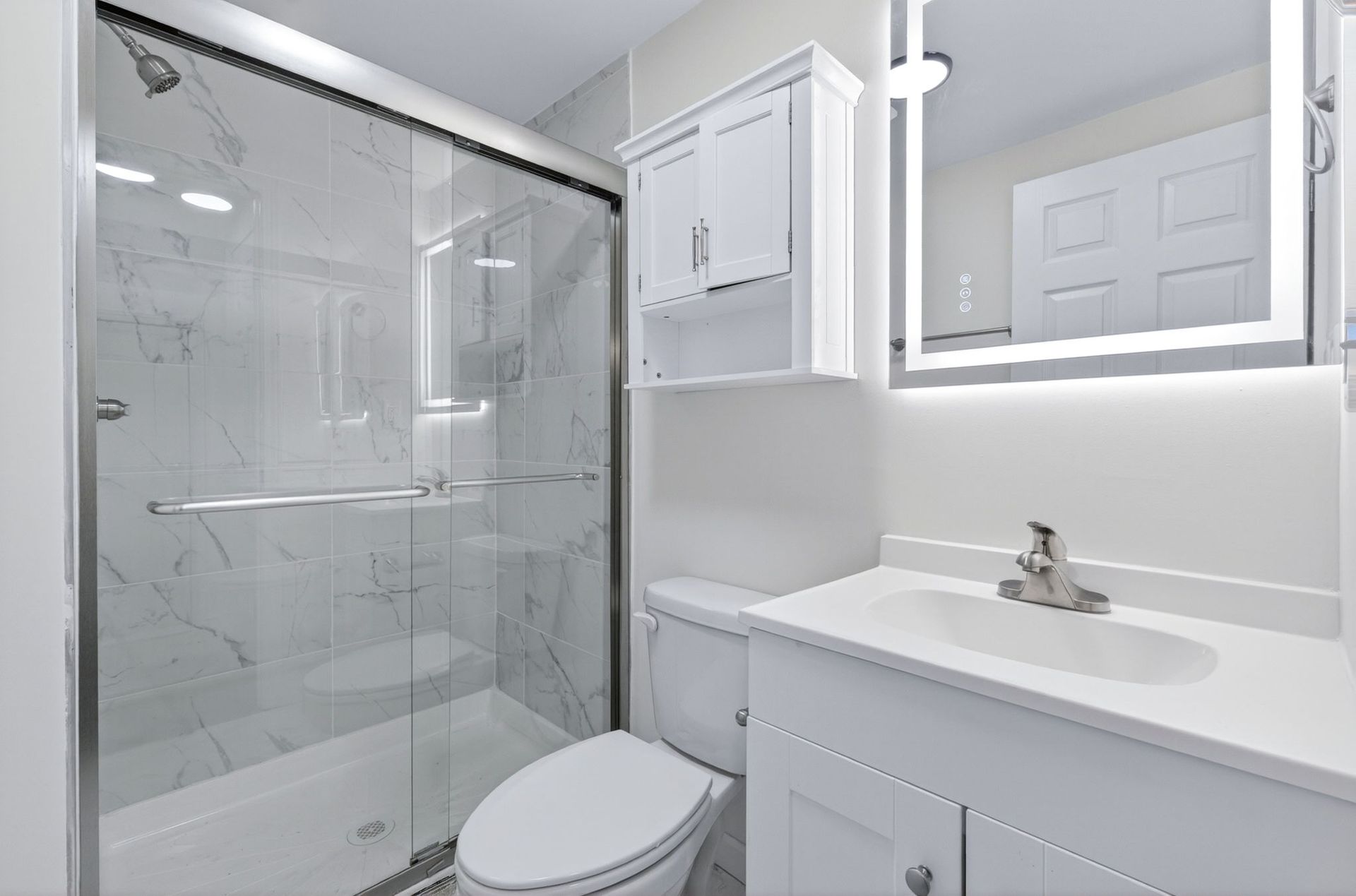 Bright white bathroom with glass shower, toilet, vanity sink, and mirrored cabinet.