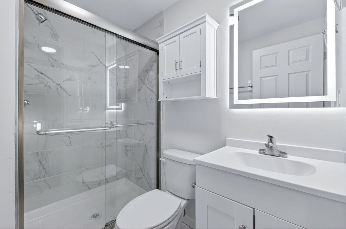 Bright white bathroom with glass shower, toilet, sink, and illuminated mirror