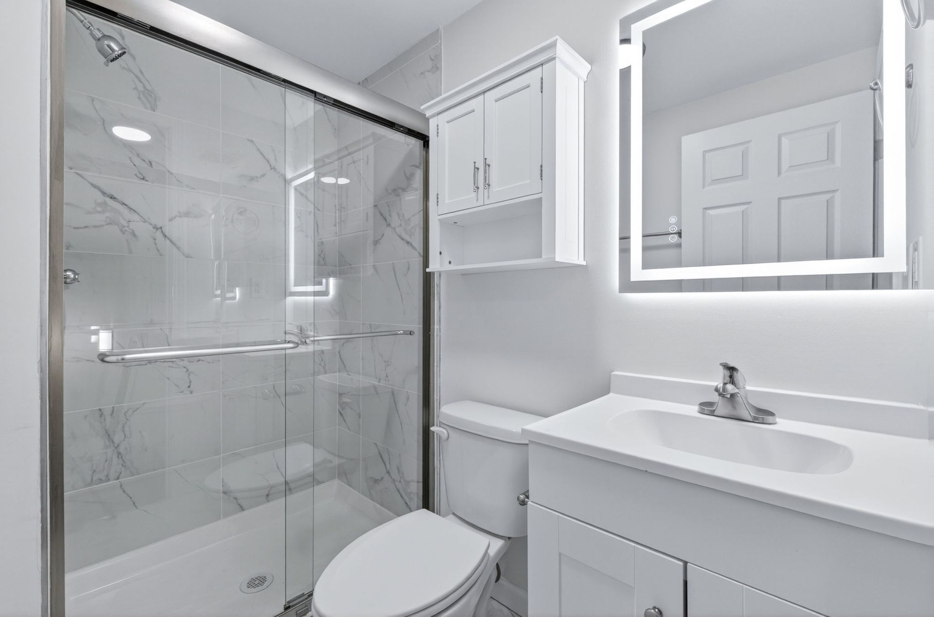 Modern white bathroom with glass shower, toilet, sink, and illuminated mirror