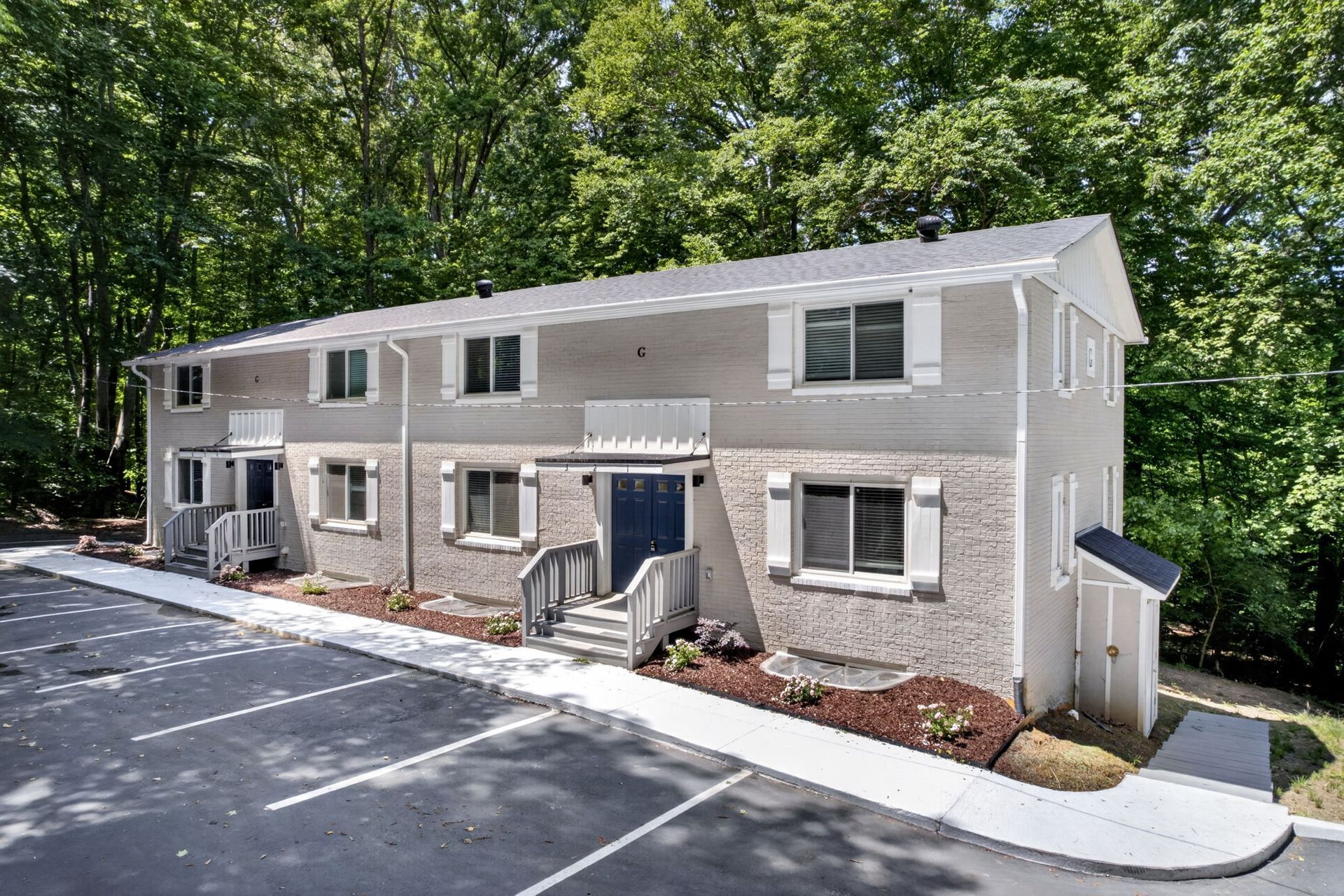 Two-story beige apartment building with white trim and parking lot in front, surrounded by trees