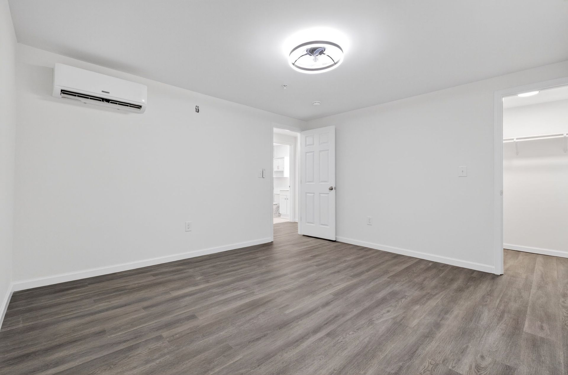 Empty white room with gray wood flooring, ceiling light, and wall-mounted air conditioner.