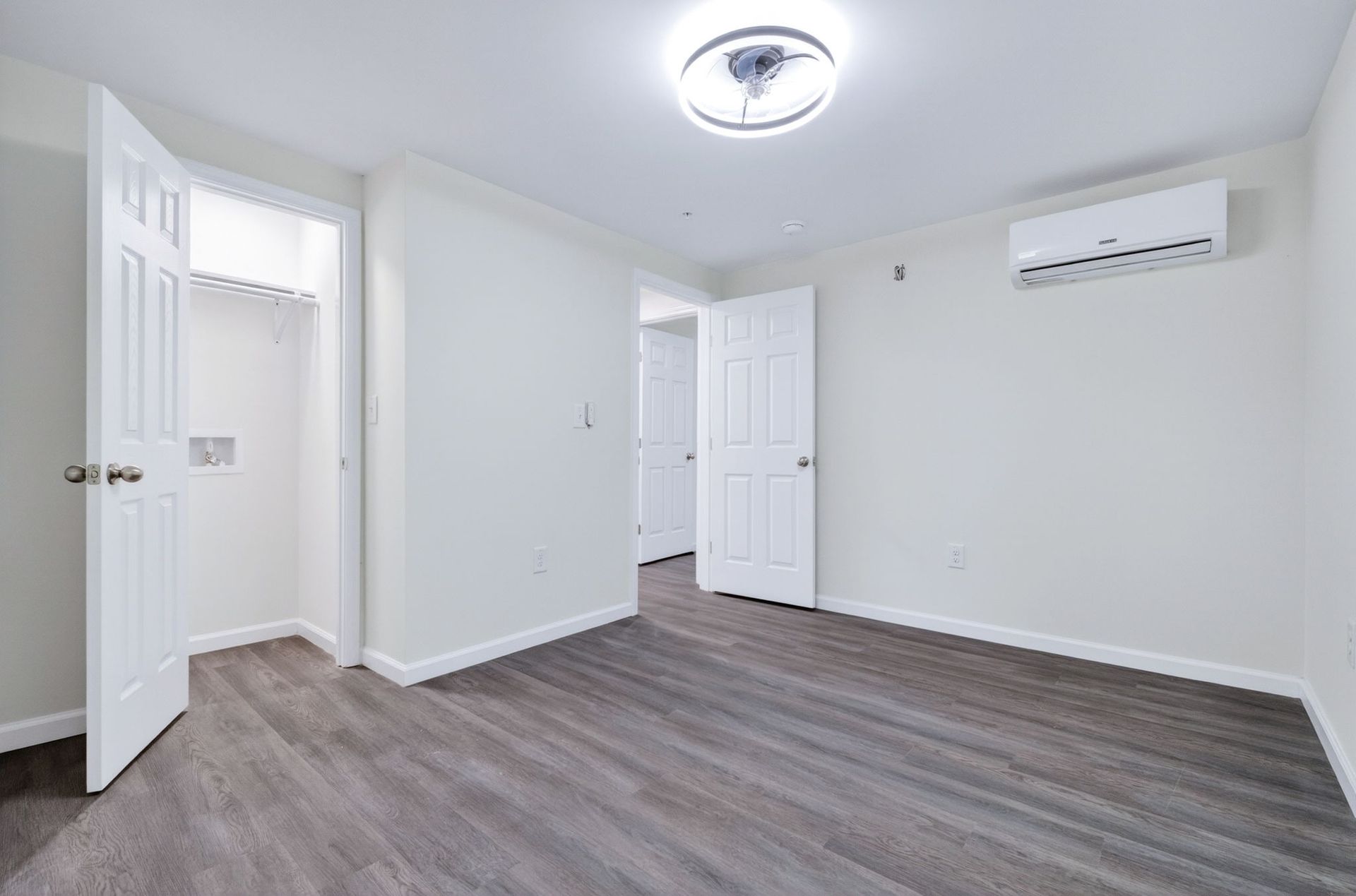 Empty white bedroom with gray wood flooring, closet doors, and a wall-mounted air conditioner.