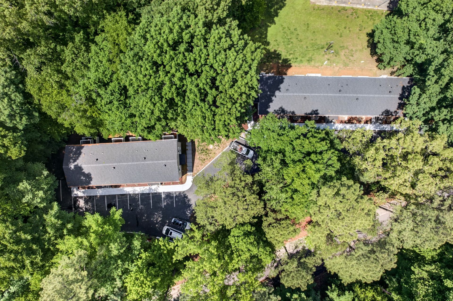 Aerial view of two houses with dark roofs surrounded by dense green trees and a driveway.