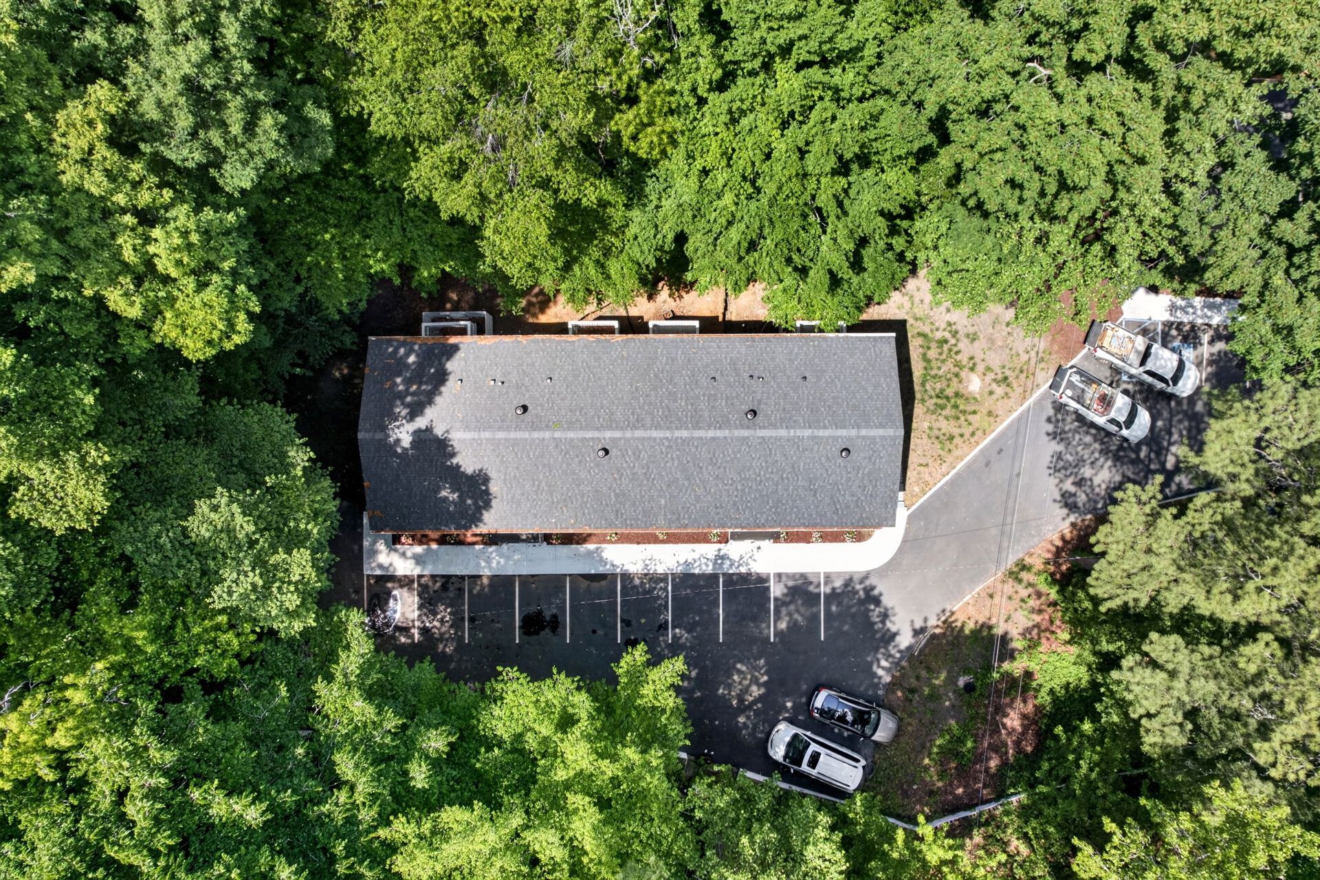 Aerial view of a house surrounded by dense green trees, with a driveway and parked vehicles.