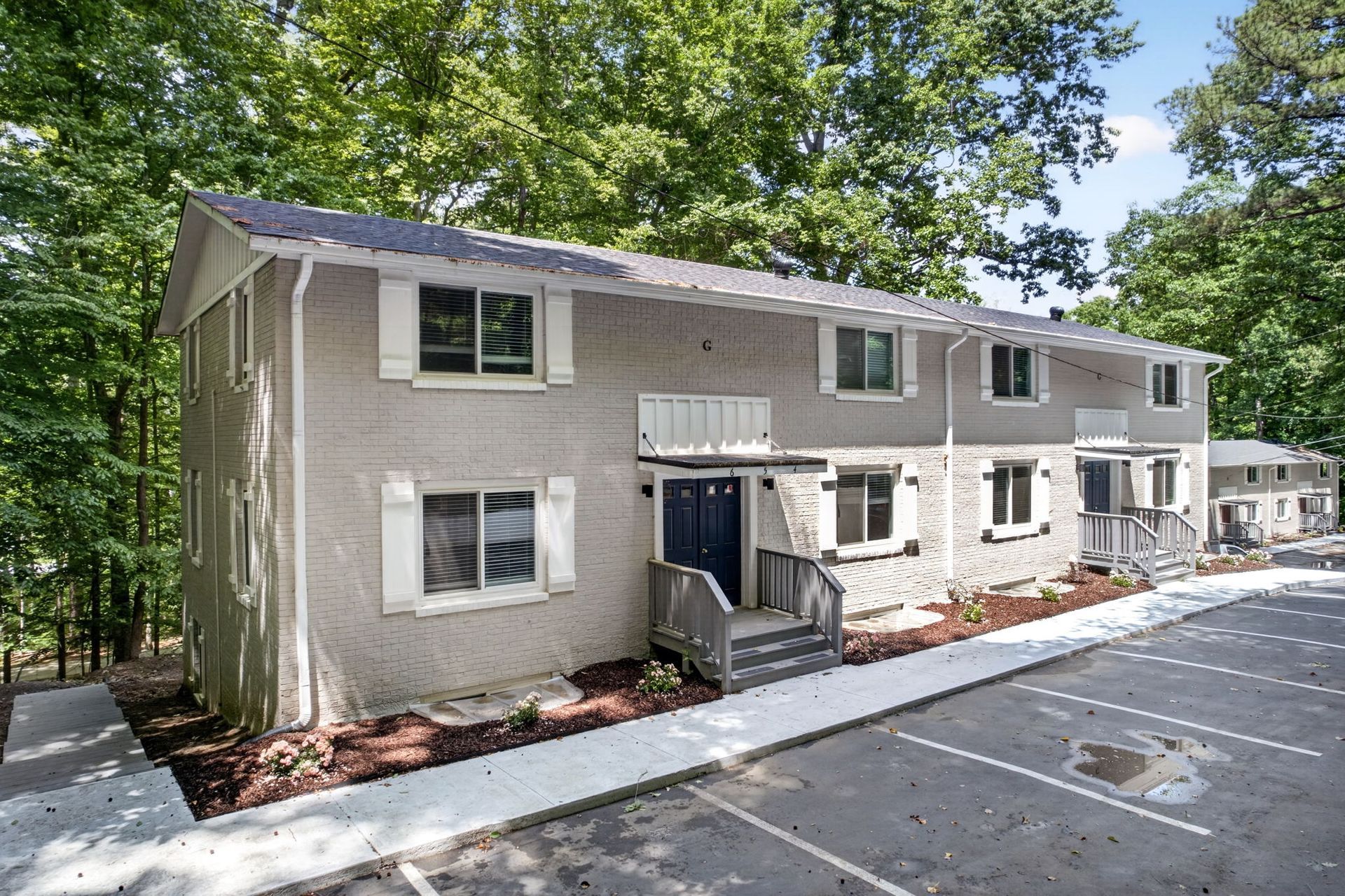 Two-story beige apartment building with white trim and a parking lot in front, surrounded by trees