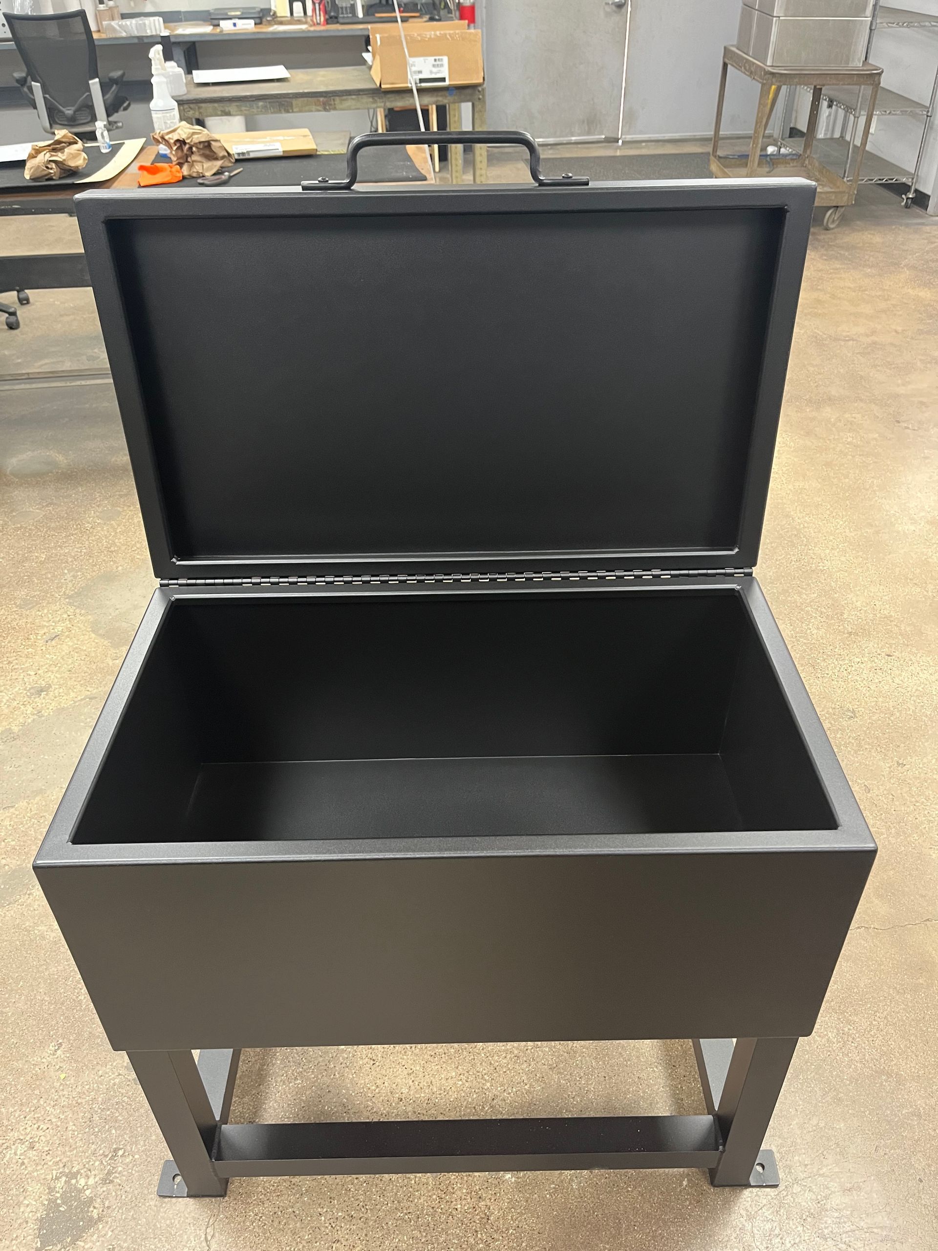 Black metal box with open lid and handle, mounted on a metal stand.