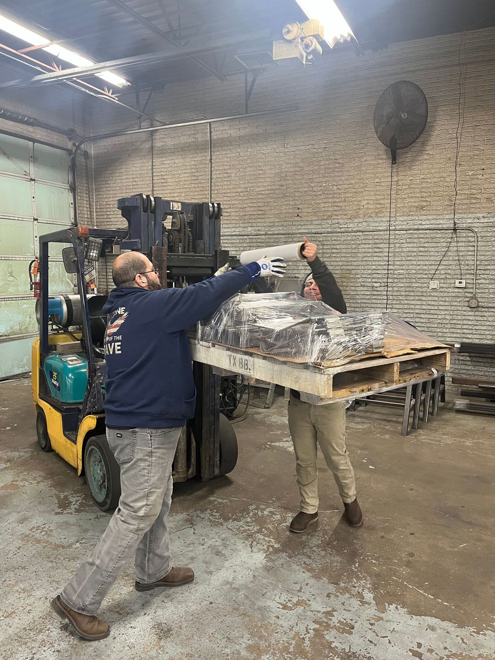 Two men loading a pallet of materials with a forklift in a warehouse; one guides, the other operates.