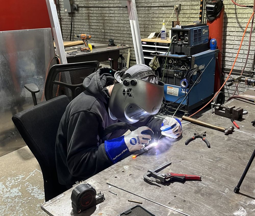 Person welding metal in a workshop, wearing a mask and gloves. Sparks fly from the welding.