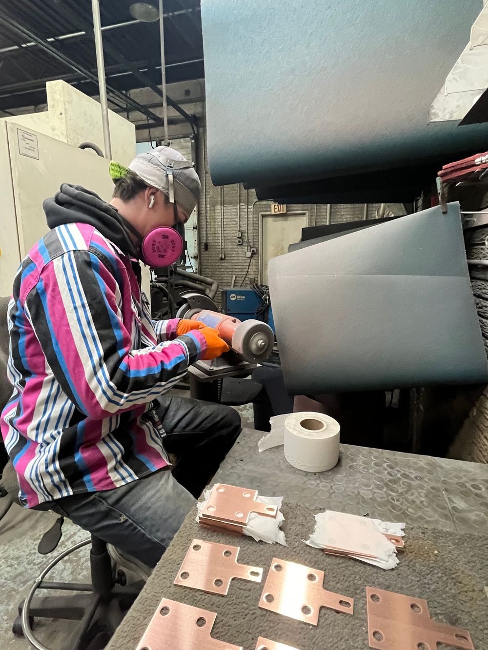 Person in mask grinds metal parts in a workshop. Pink, black, and white striped shirt, orange gloves.