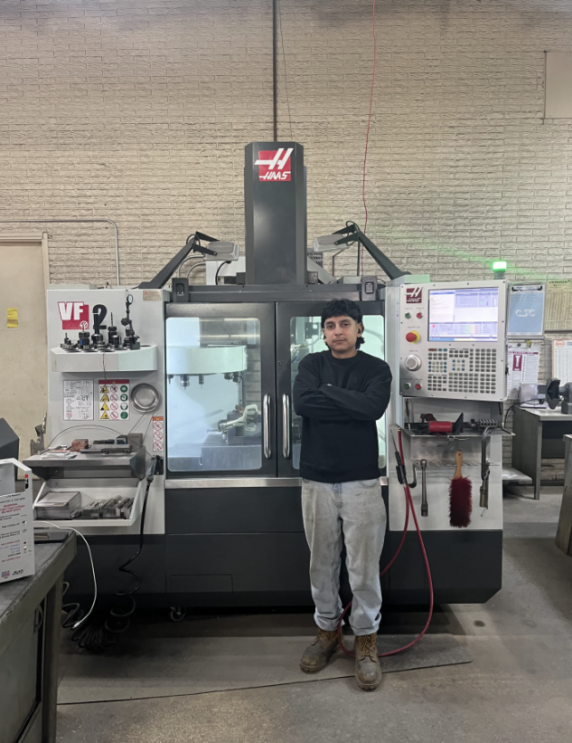A person with arms crossed stands in front of a large CNC machine in a factory setting.
