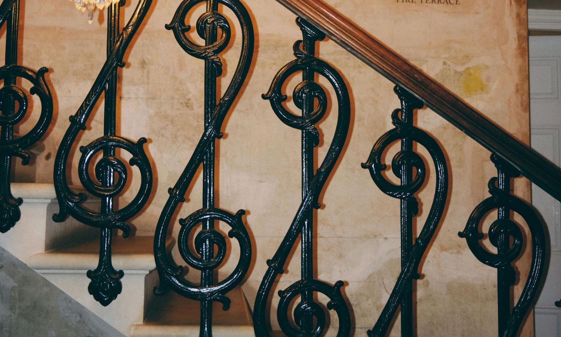 Ornate black wrought iron stair railing with scrollwork against a beige wall and wooden handrail.