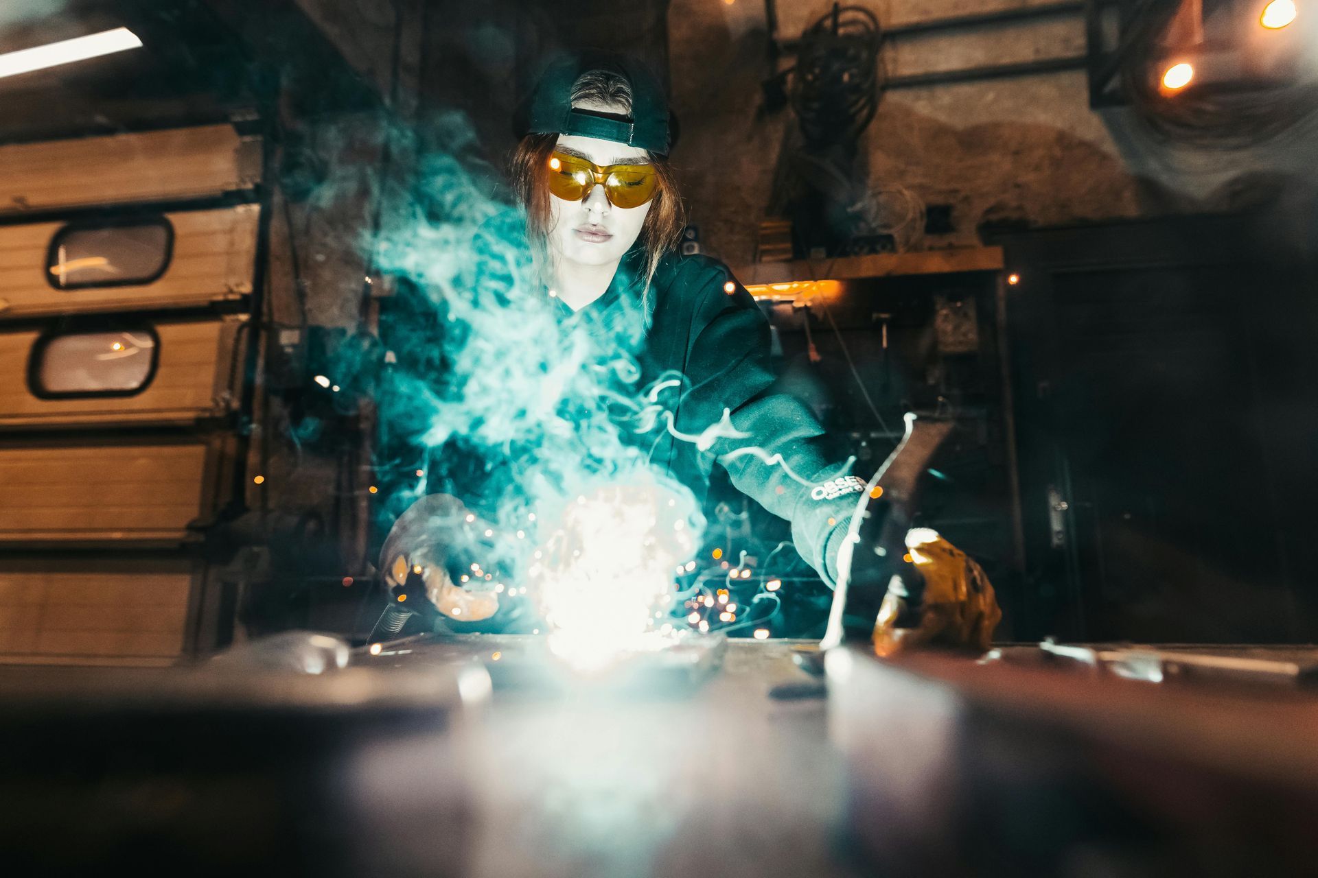 Welder in a workshop, using a welding torch. Bright blue arc, sparks, protective gear, focused expression.
