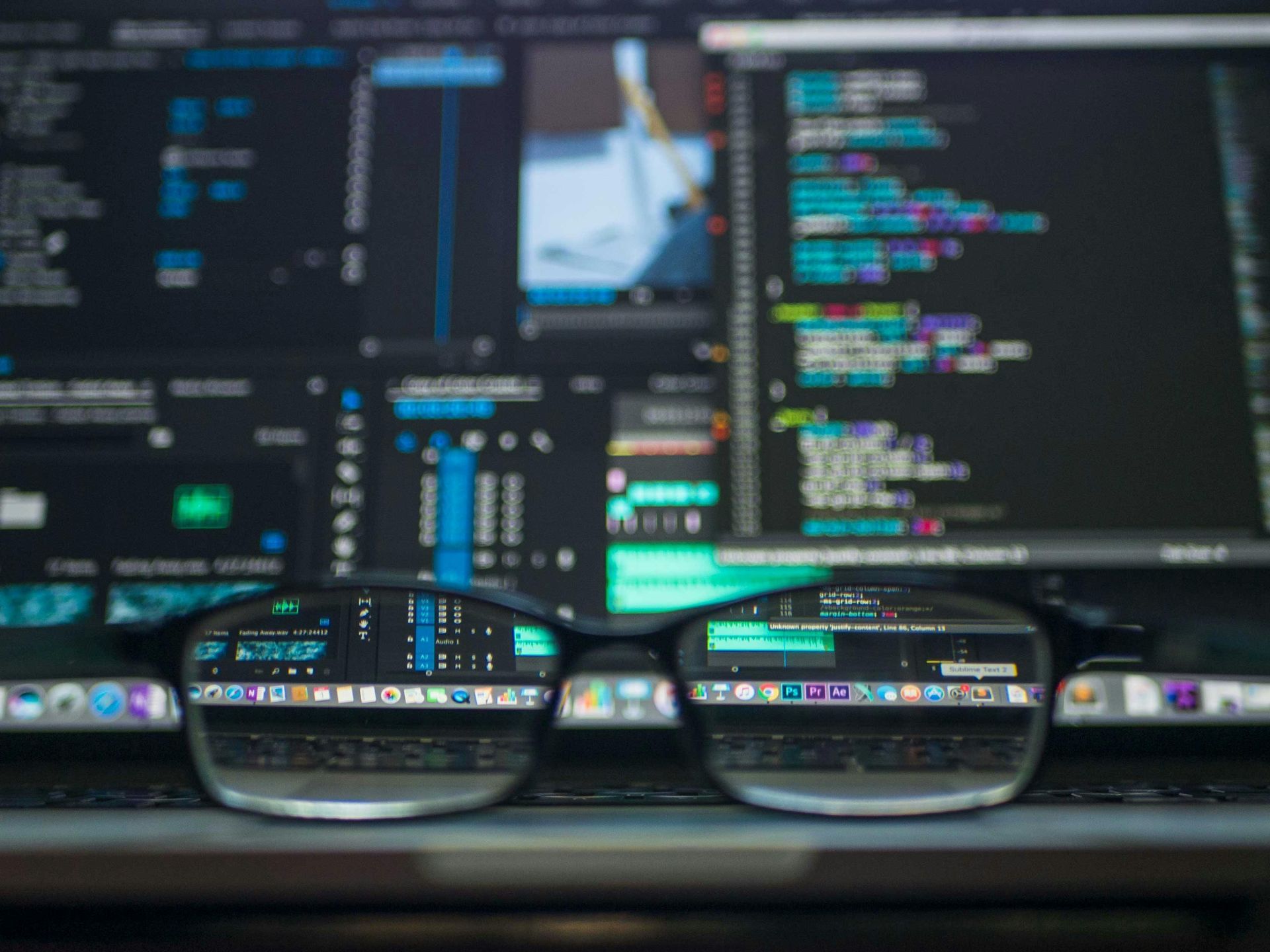 Glasses reflect a laptop screen displaying code; blurred monitor in the background.