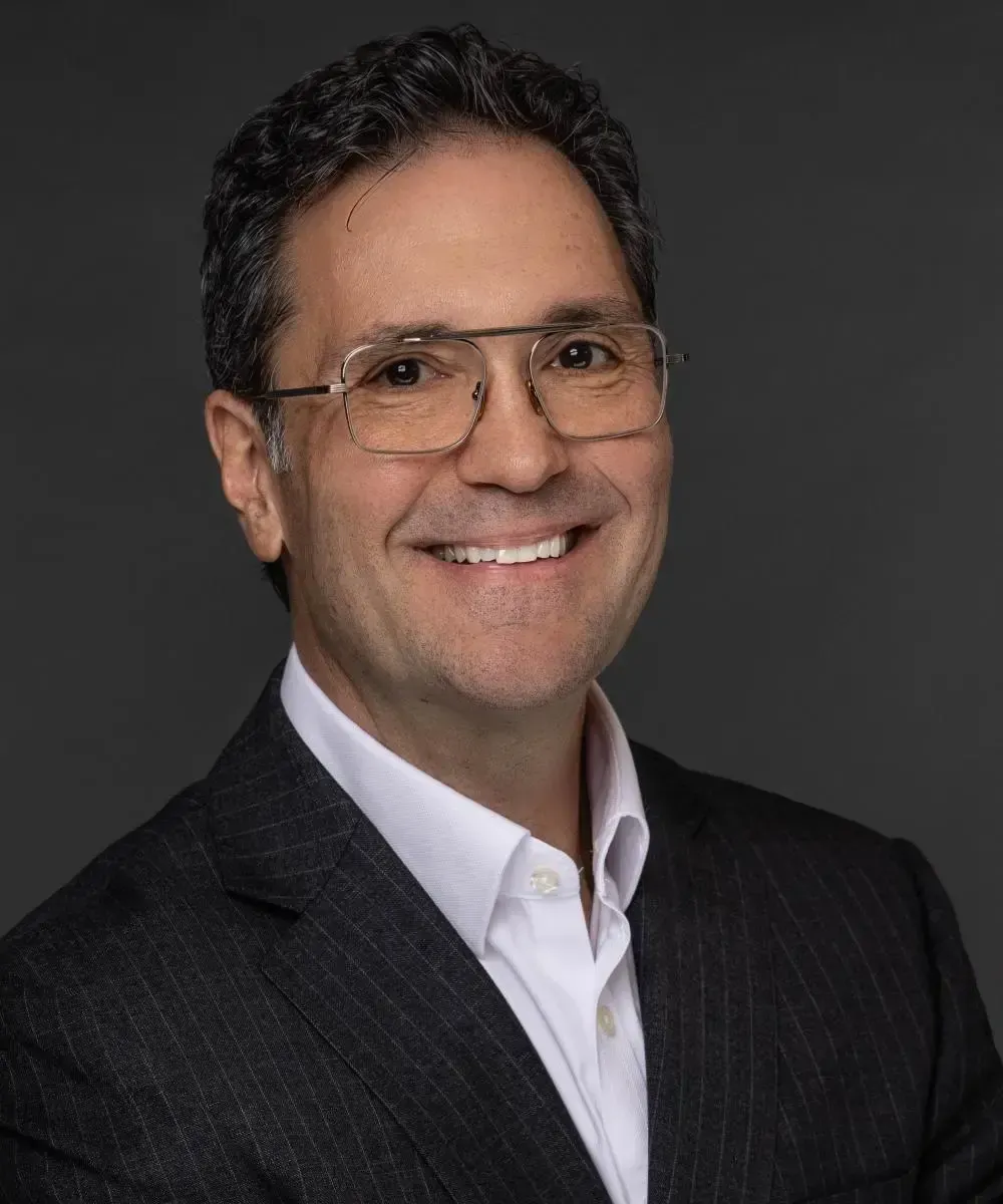 A smiling man with glasses in a dark suit and white shirt, looking towards the camera.