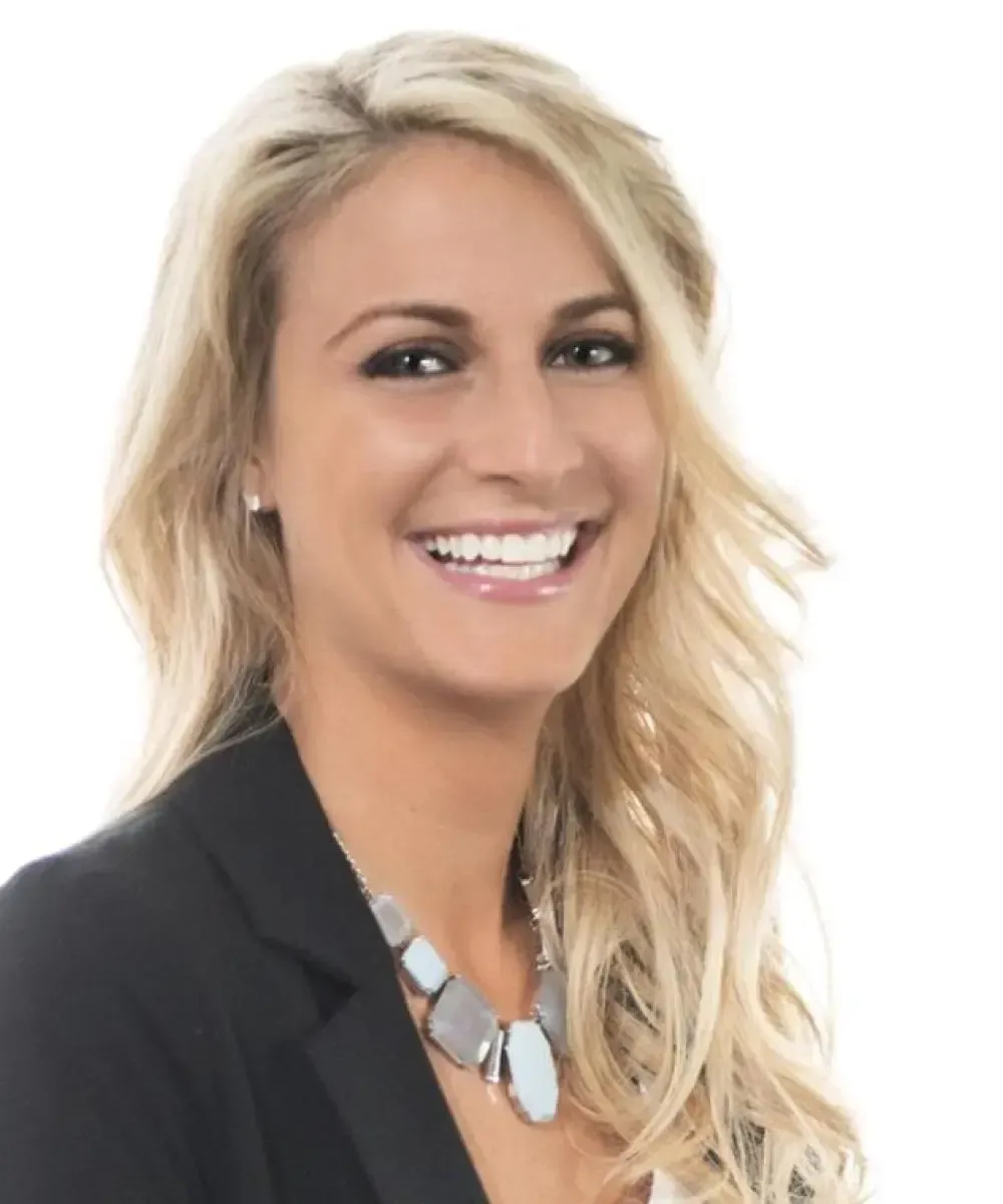 Blonde woman in a black blazer smiles broadly, wearing a statement necklace; white background.