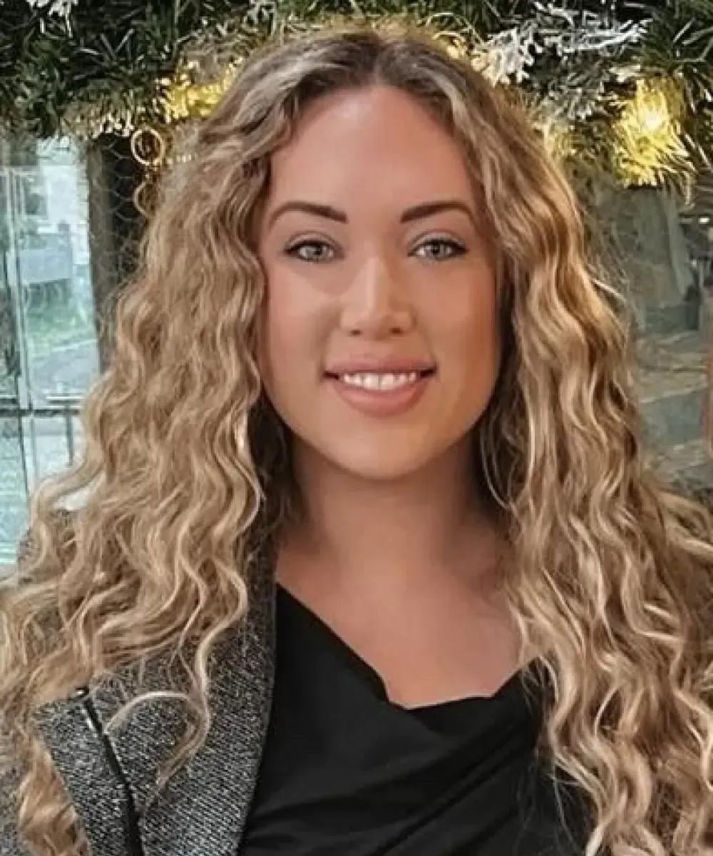 A young woman with long, curly blonde hair smiles at the camera. She wears a black top and a gray blazer.