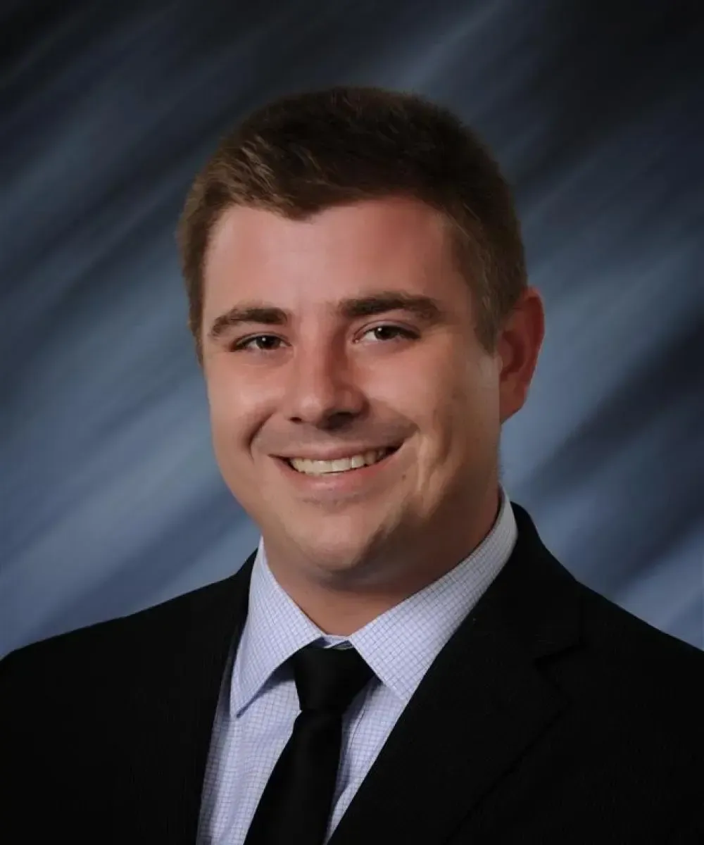 Smiling man in a suit and tie, with a blue-gray blurred background.