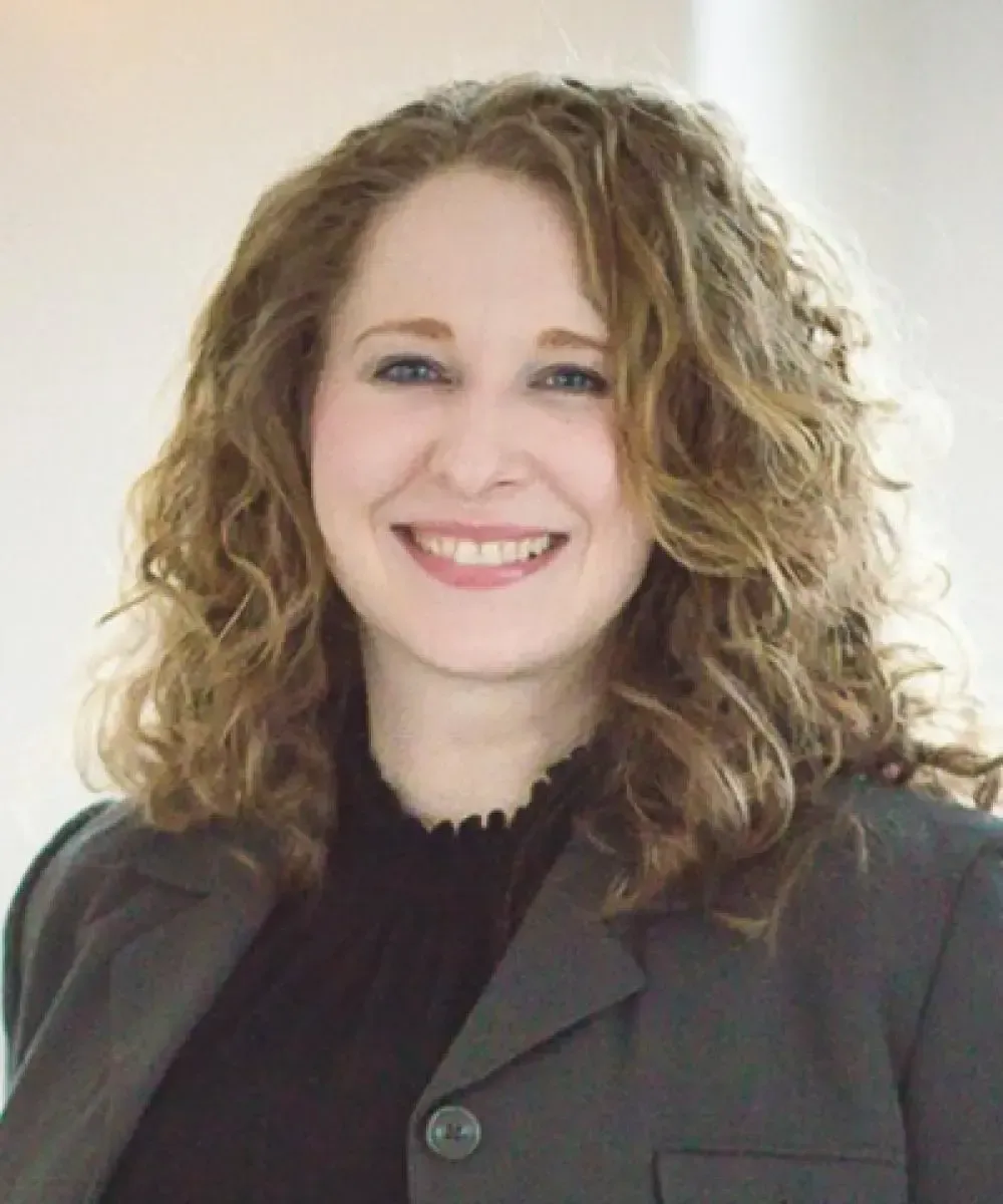 Woman with curly brown hair smiles at the camera, wearing a gray blazer and black top. Bright, neutral background.