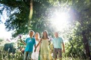 Four people walking through a sunlit wooded path, surrounded by tall grass and trees.