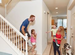Family gathered in a bright hallway beside a staircase, with a dog watching nearby.