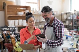 Two coworkers in aprons reviewing a tablet in a bright workshop, surrounded by pottery supplies.