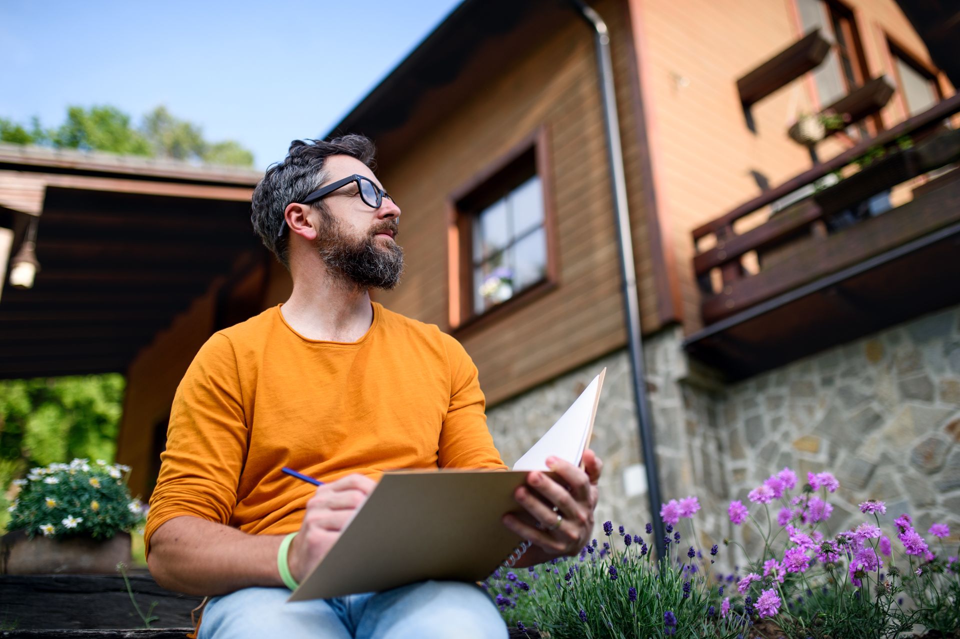 Man in orange sweater using a laptop outdoors beside purple flowers and a stone house