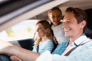 Smiling family riding in a car, with a man driving, woman in back, and child leaning forward.