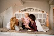 Family lounging on a bed, smiling at a baby in pink pajamas in a cozy bedroom with warm string lights