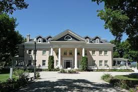 A large white house with columns and a driveway is surrounded by trees.