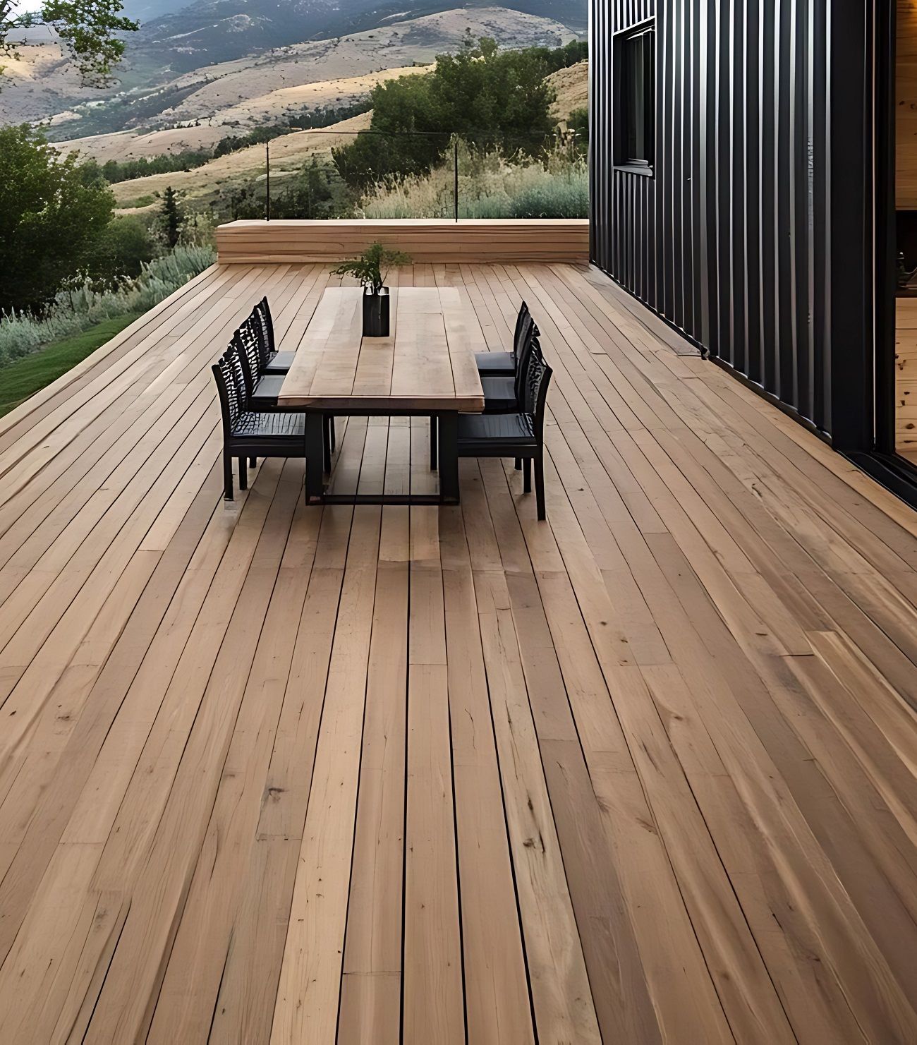 Wooden deck with a black dining table and chairs beside a modern house, overlooking a hillside view