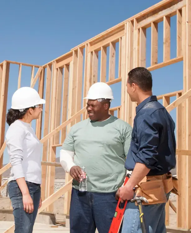 Three construction workers in hard hats discussing a framed house under construction.