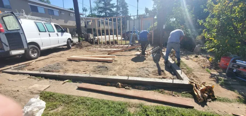 People working in a sunny yard beside a white van, moving logs and lumber near a dirt path