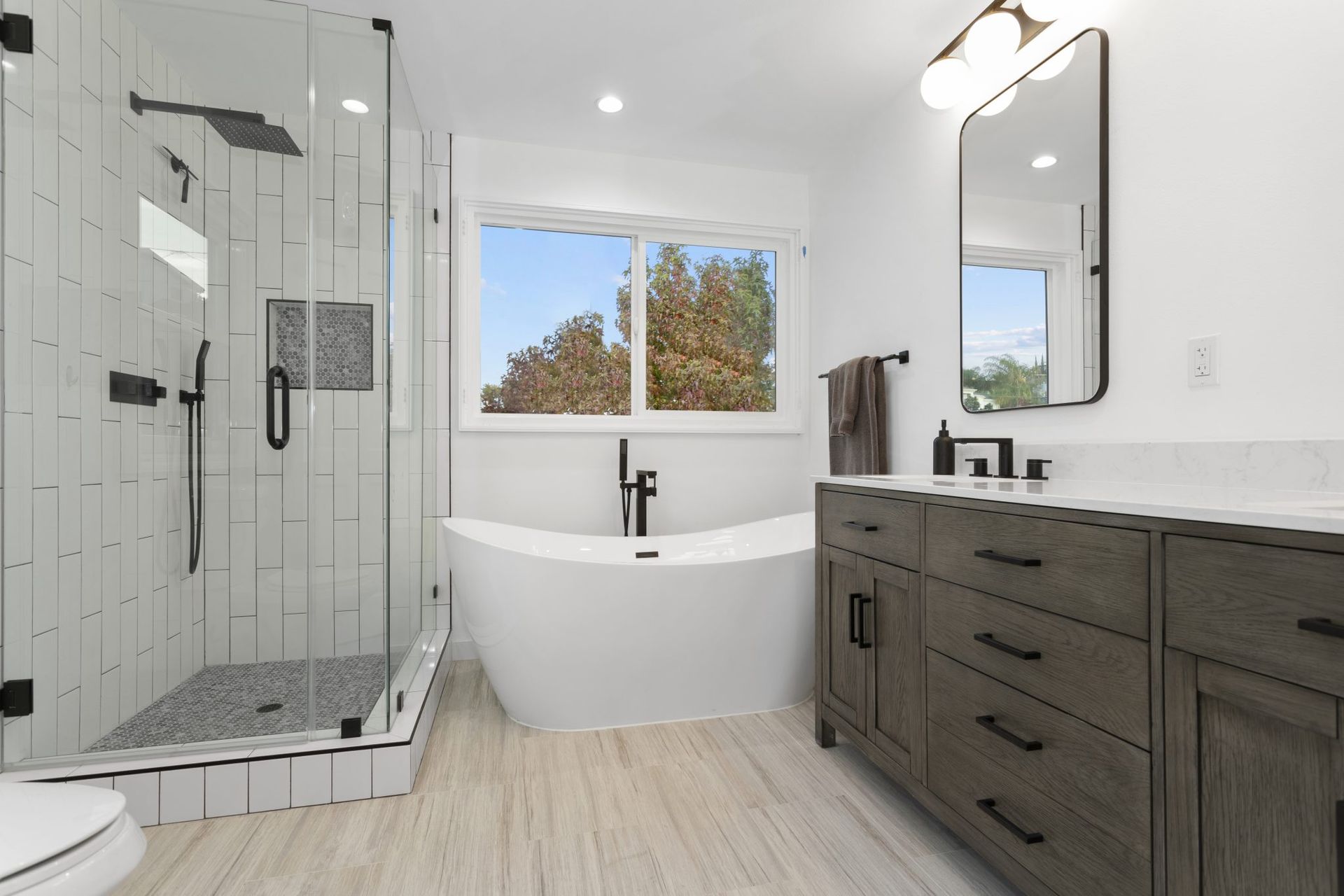 Modern bathroom with glass shower, freestanding tub, dark vanity, and window above the tub